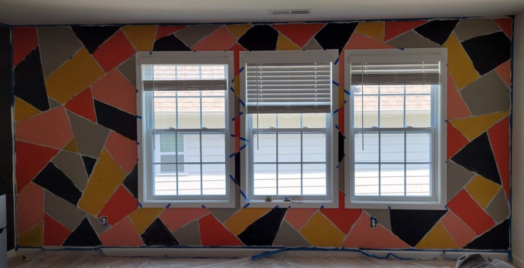 Parede interna durante processo de pintura geométrica DIY. As formas triangulares já foram preenchidas com tintas coloridas (coral, amarelo, cinza e preto), mas a fita crepe azul ainda está colada delimitando os contornos antes da remoção.