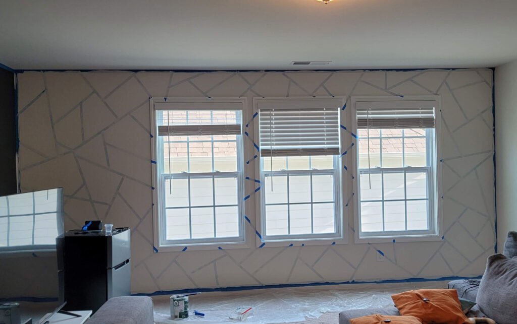 Parede preparada com fita crepe azul recebendo uma camada de tinta branca sobre as bordas da fita para selagem. Materiais de pintura como lata e rolo estão no chão protegido por lona plástica.