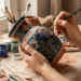 Mulher pintando à mão um vaso de cerâmica com detalhes florais em tons de azul, verde e terracota, sobre uma mesa rústica com pincéis, tintas e argila.