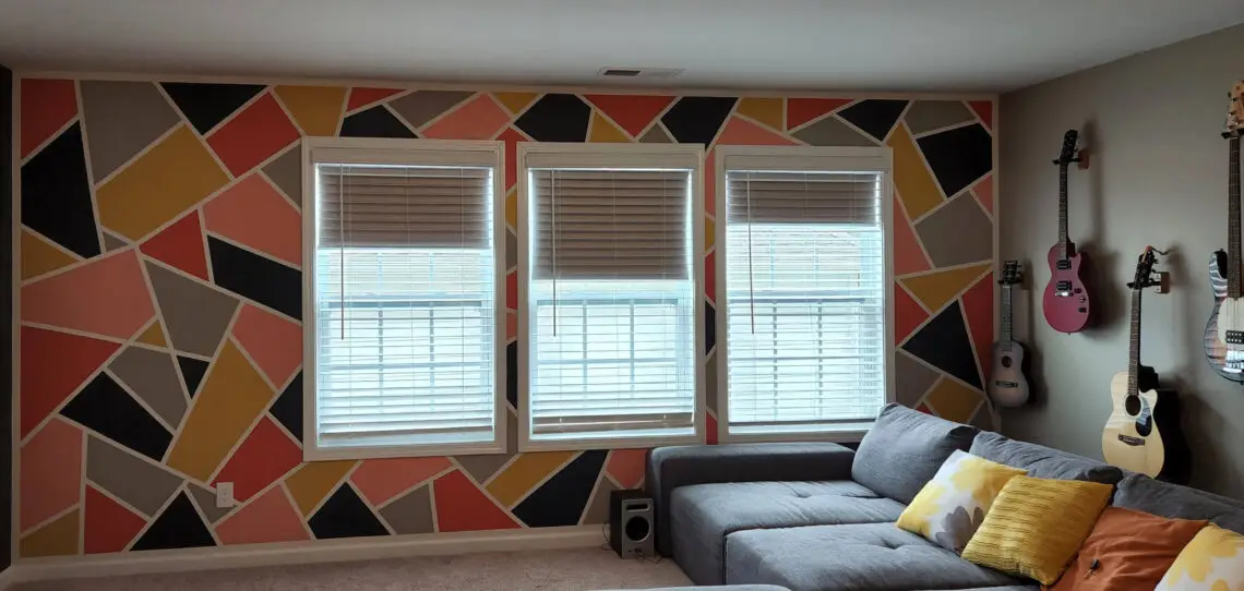 Resultado final de uma parede interna com pintura geométrica vibrante, apresentando formas triangulares e irregulares nas cores coral, amarelo mostarda, cinza escuro e preto, separadas por linhas brancas. A pintura contorna três janelas centrais com persianas brancas.