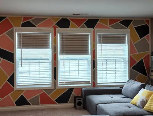 Resultado final de uma parede interna com pintura geométrica vibrante, apresentando formas triangulares e irregulares nas cores coral, amarelo mostarda, cinza escuro e preto, separadas por linhas brancas. A pintura contorna três janelas centrais com persianas brancas.