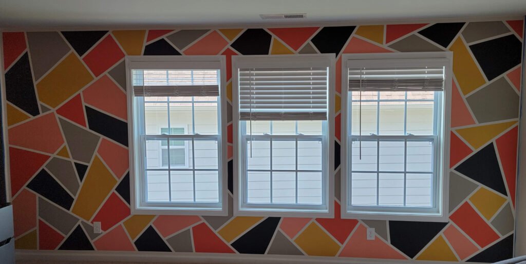 Resultado final de uma parede interna com pintura geométrica vibrante, apresentando formas triangulares e irregulares nas cores coral, amarelo mostarda, cinza escuro e preto, separadas por linhas brancas. A pintura contorna três janelas centrais com persianas brancas.