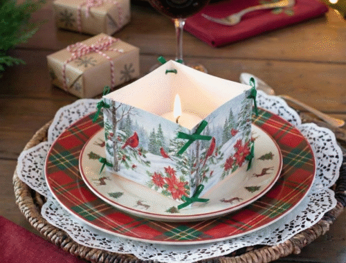 Mesa de Natal rústica posta com um centro de mesa quadrado de papel ilustrado com cardeais e vela acesa, sobre pratos xadrez vermelhos e verdes. Uma taça com vinho tinto, presentes embrulhados em papel pardo e um fundo com luzes de árvore de Natal desfocadas completam a cena.