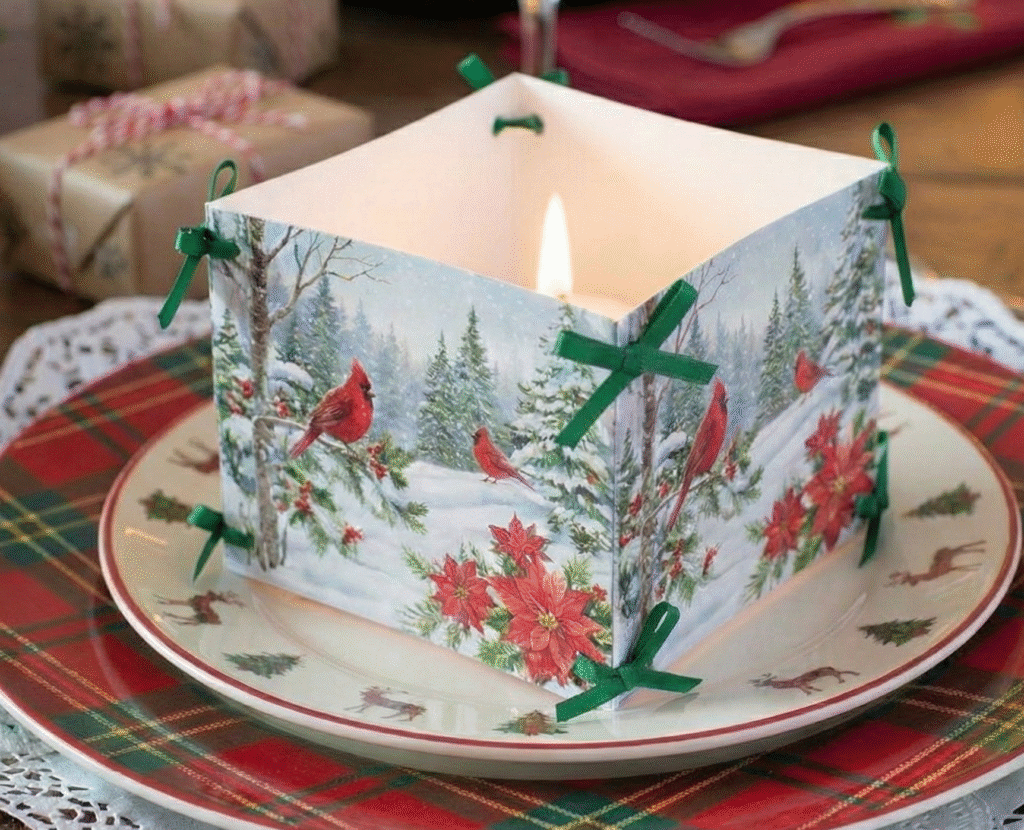 Close-up detalhado de um centro de mesa de papel quadrado com ilustração de inverno e cardeais vermelhos. Uma vela acesa dentro do cubo ilumina o papel, com laços verdes nos cantos. Os pratos xadrez estão desfocados abaixo.